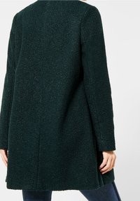 Cappotto verde scuro realizzato in tessuto testurizzato, con una vestibilità ampia, maniche lunghe e un taglio dritto con fessure laterali per agevolare i movimenti.