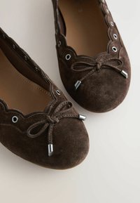 Ballerines en daim brun foncé avec bords ondulés, œillets métalliques et nœuds décoratifs noués avec embouts métalliques sur fond beige.
