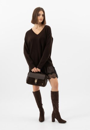 Frau trägt schwarzen Pullover mit V-Ausschnitt, schwarzen Spitzenrock und kniehohe braune Stiefel, hält eine dunkelbraune Handtasche vor weißem Hintergrund.