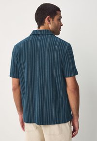 Short-sleeve, navy shirt featuring a textured vertical stripe pattern and a collared neckline, paired with light-colored pants.