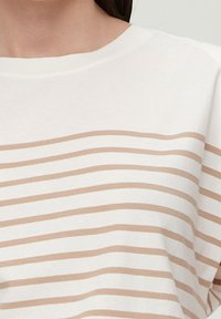 Bílá košile s světle hnědými horizontálními pruhy, kterou nosí osoba s tmavými vlasy, zkrácená tak, aby ukazovala pouze ramena a hrudník.