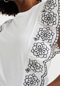 Vit T-shirt med svarta broderade blommönster och volanger på ärmarna. Slät tyg med en figurnära passform.