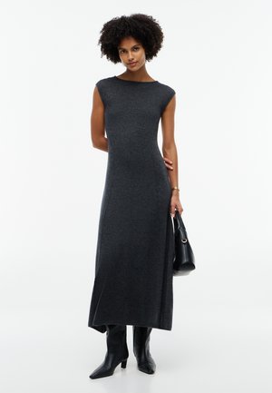 Woman with curly hair wearing a sleeveless dark gray long dress, black boots, holding a black handbag, standing against a white background.