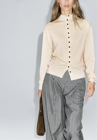 Haut beige à manches longues avec des boutons, un col montant et des boutons noirs, associé à un pantalon large rayé gris et un sac marron.