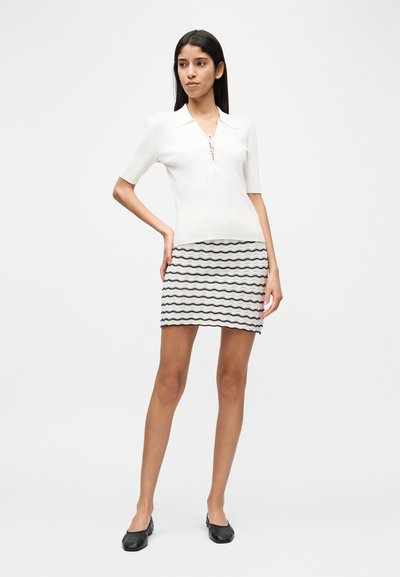 Λευκό ριμπ πουκάμισο πόλο με δέσιμο στο μπροστινό μέρος, σε συνδυασμό με μια κοντή φούστα με μαύρα κυματιστά σχέδια και μαύρα επίπεδα παπούτσια.