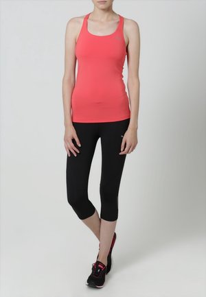 Donna in piedi che indossa una maglietta atletica senza maniche color corallo, leggings neri fino al polpaccio e scarpe da corsa nere con dettagli rosa.