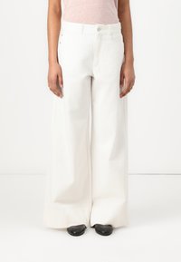 Pantaloni bianchi in cotone a gamba larga con chiusura a bottone, tasche frontali e dettagli di cucitura minimi. Abbinati a scarpe nere.