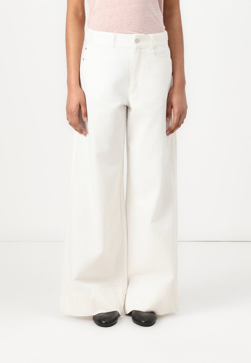 Pantaloni bianchi in cotone a gamba larga con chiusura a bottone, tasche frontali e dettagli di cucitura minimi. Abbinati a scarpe nere.
