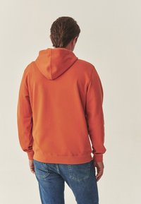Oranje hoodie met een kangoeroezak, geribde manchetten en zoom. Gemaakt van een katoenmengsel, gedragen met blauwe spijkerbroek.