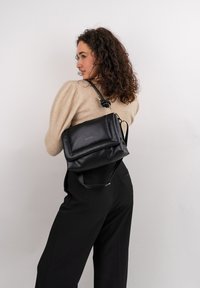 Černá kožená crossbody kabelka s designem na překlopení, zlatými kovovými doplňky a nastavitelným popruhem, nošená přes béžový top.