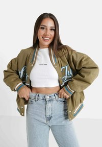 Glimlachende vrouw met lang bruin haar, gekleed in een kaki varsityjack over een witte crop top en lichtblauwe high-waisted jeans, met haar handen in haar zakken.