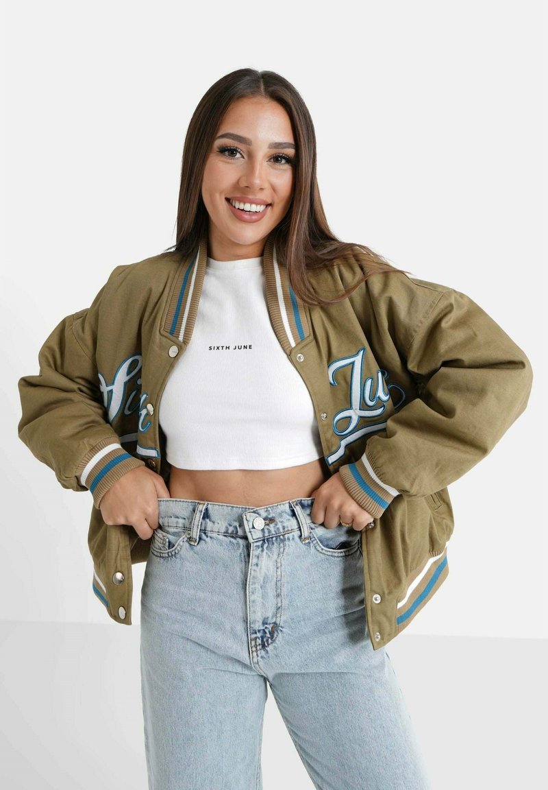 Glimlachende vrouw met lang bruin haar, gekleed in een kaki varsityjack over een witte crop top en lichtblauwe high-waisted jeans, met haar handen in haar zakken.
