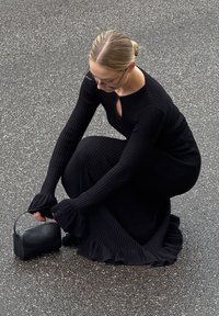 Robe noire à rayures avec des manches longues et un décolleté en trou de serrure, associée à un petit sac à main noir. Texture lisse, design ajusté, longueur au genou.