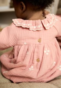 Vestito in mussola rosa con collo arricciato, ricamo floreale e bottoni in legno sul retro. Texture morbida con design a gonna arricciata.