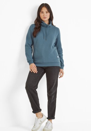 Blue fleece hoodie with a high collar and adjustable drawstrings, paired with dark jeans and white sneakers, featuring rolled cuffs.