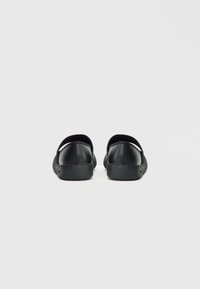 Zapatos negros de tipo slip-on con acabado de cuero liso, bordes curvados, parte trasera abierta y detalle sutil de costuras a lo largo de la suela.