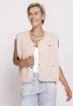 Smiling woman with short blonde hair wearing a beige fleece vest over a white top and light blue denim jeans, standing against a light background.