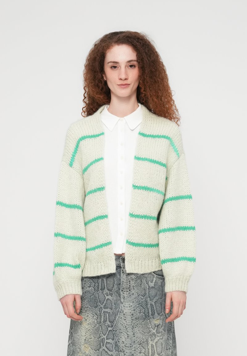 Νεαρή γυναίκα με κατσαρά κόκκινα μαλλιά, που φοράει λευκό πουκάμισο με κουμπιά, μπεζ ζακέτα με πράσινες ρίγες και γκρι φούστα με μοτίβο φίδι.