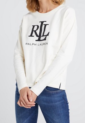 Kobieta w białej, długorękawowej bluzie Ralph Lauren z czarnym logo i niebieskich dżinsach, z dłońmi splecionymi przed sobą, na jednolitym tle.