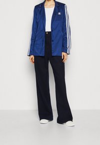 Tmavě modrý saténový blejzr s bílými třemi pruhy, dvěma knoflíky a logem Adidas. Kombinován s tmavými kalhotami s vysokým pasem a bílými botami.