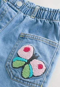 Poche de pantalon en denim bleu avec un écusson papillon texturé et coloré présentant des détails roses, verts, turquoise et rouges.