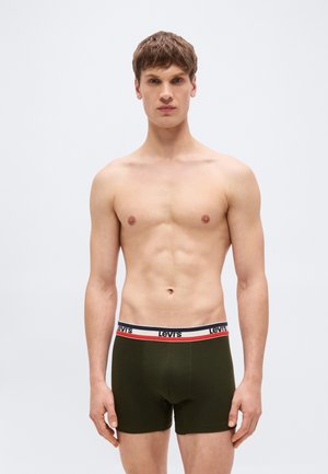 Νέος άνδρας με κοντά καστανά μαλλιά που φοράει σκούρα πράσινα εσώρουχα Levi's τύπου μποξέρ με λογότυπο στη μέση, στέκεται μπροστά σε απλό ανοιχτό φόντο.