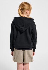 Schwarzer Kapuzenpullover mit texturiertem Finish, versehen mit seitlichen Akzenten. Kombiniert mit hellbeigen Shorts, von hinten gesehen.