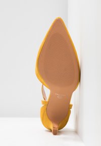 Chaussure en daim jaune à bout pointu avec petit talon, vue de la semelle avec une semelle ondulée et bride à la cheville sur fond blanc.