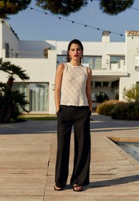 Beige strikket tanktop med et struktureret m�ønster, parret med skræddersyede sorte brede bukser og sorte sandaler. Baggrunden indeholder grønne planter og arkitektur.