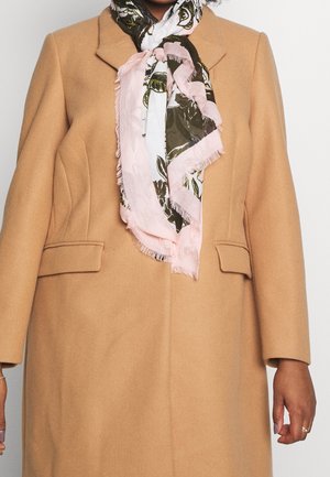 Femme portant un manteau couleur chameau avec des poches à rabat et une écharpe à motifs floraux rose et olive nouée autour du cou.