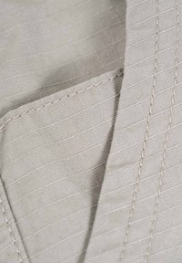 Hellbeige Stoff mit einem strukturierten Muster, das subtile diagonale Linien und sichtbare Nähte entlang der Ränder aufweist. Glatte Oberflächenbeschaffenheit.