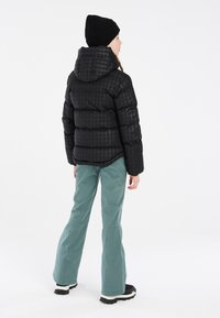Giacca a vento nera con cappuccio e texture a motivi, abbinata a pantaloni a campana color teal e scarpe nere, vista da dietro.