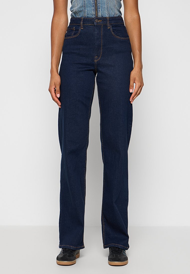 Dunkelblaue Denim-Jeans mit hoher Taille, geradem Bein, kontrastierenden orangefarbenen Nähten und Fronttaschen. Getragen mit schwarzen Schuhen.