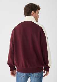 Felpa marrone con dettagli crema, polsini e orlo a coste, texture morbida e collo alto. Presenta una vestibilità rilassata e un design a tinta unita.