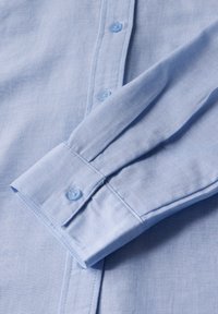 Camicia azzurra chiaro con tessuto leggermente strutturato. Presenta bottoni azzurri coordinati e un polsino con cuciture di dettaglio.