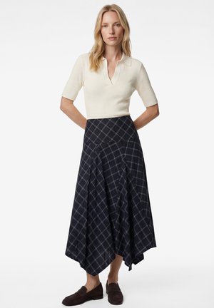 Kobieta stojąca z rękami z tyłu, ubrana w kremowy krótki sweter z krótkim rękawem, granatową spódnicę w kratę oraz ciemnobrązowe mokasyny.