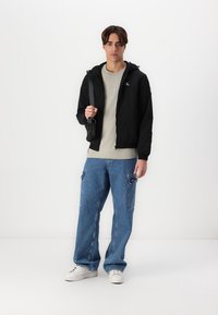 Črna jakna s kapuco, siva majica s logotipom, svetlo modri široki jeans, beli superge, drži majhno črno torbico.
