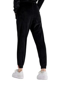 Schwarze Velour-Jogginghosen mit elastischen Bündchen, ausgestattet mit einem Kordelzugbund und einem kleinen roten Logo-Detail auf der Rückseite. Kombiniert mit weißen Sneakers.