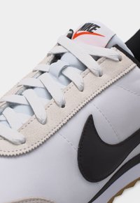 Λευκό αθλητικό παπούτσι Nike με μαύρο λογότυπο swoosh, λευκά κορδόνια, μπεζ σουέντ διακοσμητικά και λευκή γλώσσα με μαύρο και πορτοκαλί λογότυπο Nike.