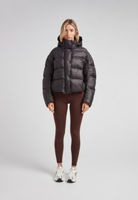 Donna bionda che indossa un giubbotto a vento nero, leggings marroni e sneakers bianche, in piedi contro uno sfondo grigio chiaro uniforme.