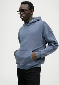 Sweatshirt azul feito de um tecido macio, com um cordão, bolso canguru e punhos canelados. Usado com calças escuras, o modelo está posicionado contra um fundo neutro.