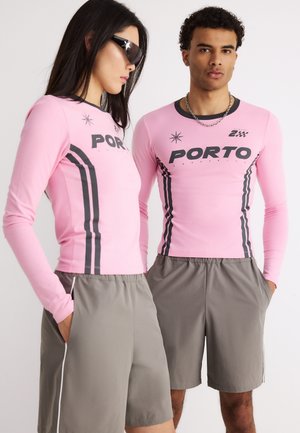 Mężczyzna i kobieta w pasujących różowych bluzkach z długim rękawem z napisem "PORTO" i szarych szortach, stojący z rękami w kieszeniach na białym tle.