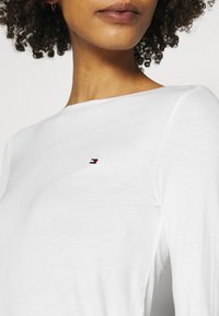 Camiseta blanca de manga larga con cuello redondo y sutil logo de Tommy Hilfiger en azul marino, rojo y blanco en la parte izquierda del pecho. Textura suave.
