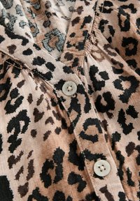 Blůza s leopardím potiskem ve světle hnědé a černé barvě. Obsahuje zapínání na knoflíky, texturovanou látku a skladbu v okolí výstřihu.