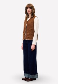 Frau in brauner Knopfveste über einem weißen Langarmshirt, mit weit geschnittenen dunkelblauen Jeans und hochgekrempelten Hosenbeinen sowie braunen Lederschuhen, steht seitlich.