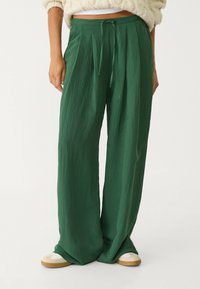 Persoon in hoog-taille, wijdvallende donkergroene broek met een koord in de taille en crèmekleurige sneakers, staande tegen een effen achtergrond.