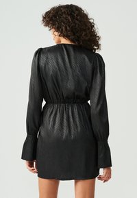 Zwarte jurk van zijdeachtig materiaal met een textuurpatroon, lange mouwen, elastische taille en klokmouwen. Achteraanzicht toont de halslijn en pasvorm.