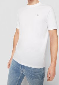 T-shirt bianco in cotone con maniche corte e colletto rotondo, con un piccolo logo nero sul petto. Indossato con jeans azzurri chiari.