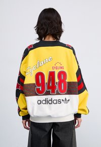 Kleurige oversized sweatshirt met een gele basis, zwarte accenten en rode sterpatronen. Bevat tekst: "Cyclone 948" en het "adidas" logo.