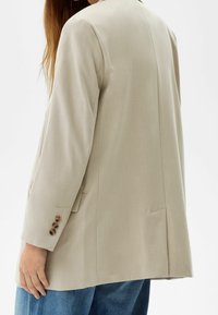 Blazer beige dal taglio rilassato, con spacco singolo, polsini abbottonati e tessuto liscio. Indossato sopra jeans blu, che evidenziano un design minimal.